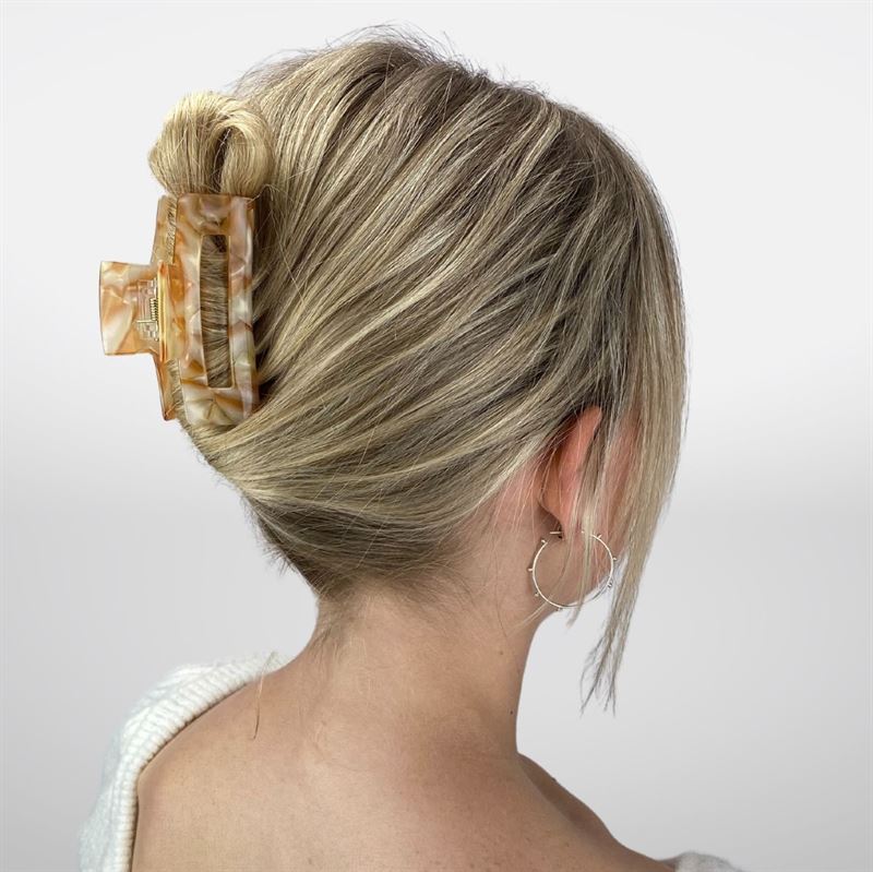 A person with blonde hair in a French twist, secured by the SOHO Aria Hair Claw in Champagne—a large, marbled accessory from SOHO. Seen from behind, they wear a white top and silver hoops; the stylish hair clip completes their look.