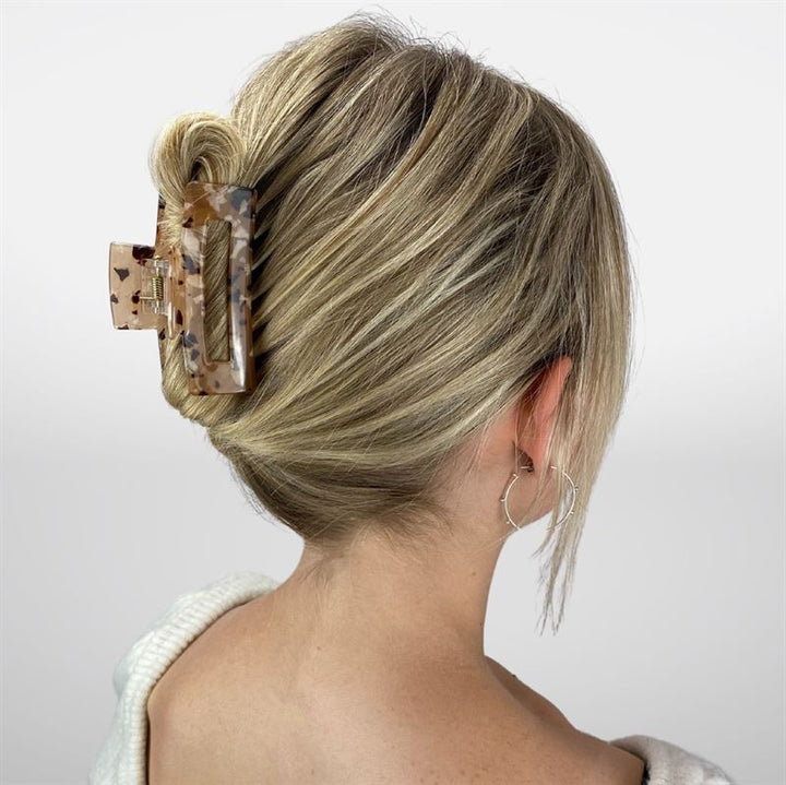 Eine Frau mit dichtem blondem Haar, das zu einer niedrigen Flechte gestylt und mit der SOHO Aria Hair Claw in Light Tortoise befestigt ist, trägt silberne Creolen und ein weißes Oberteil und steht mit dem Rücken zur Kamera vor einem einfarbigen Hintergrund.