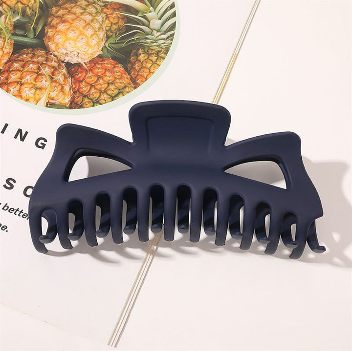 The SOHO Astrid Hair Claw in Navy by SOHO sits on an open book, with a glimpse of pineapple in the background—an ideal, stylish hair accessory for thick hair that offers a secure hold.