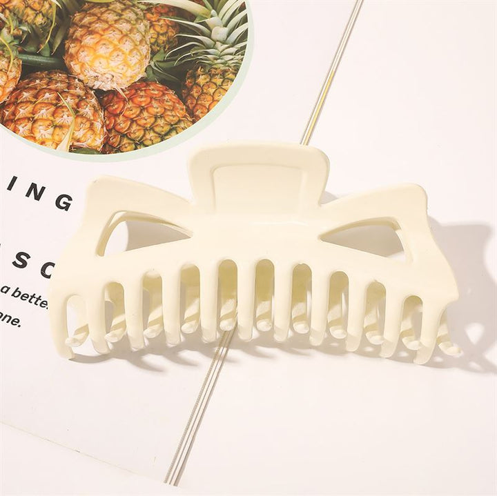 The SOHO Astrid Hair Claw - White by SOHO, designed for thick hair, rests on an open magazine page with a pineapple photo in the corner, casting a soft shadow on the white background.