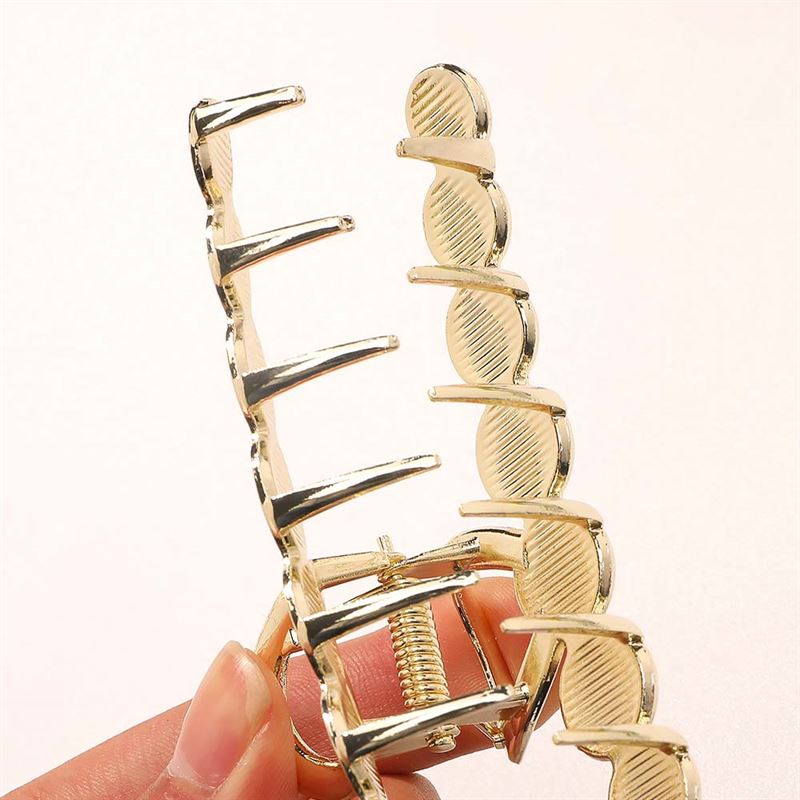 Nahaufnahme einer Hand, die die SOHO Becca Hair Claw – Midnight von SOHO hält, mit einem wellenförmigen, goldfarbenen Metalldesign und ineinandergreifenden Zinken für einen sicheren Messy Bun, vor einem einfarbigen hellen Hintergrund.