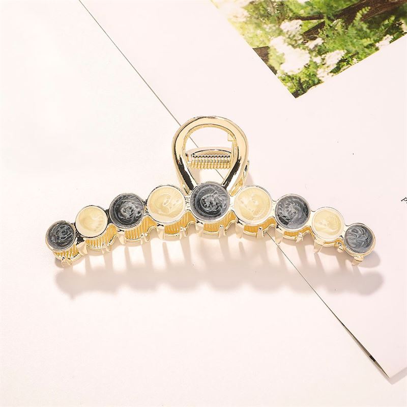 Die SOHO Becca Hair Claw – Midnight von SOHO besteht aus goldenem Metall mit schwarzen und cremefarbenen Akzenten und wird auf einer weißen Oberfläche in der Nähe von Blumenpapier gezeigt – ideal, um unordentliche Dutts zu fixieren oder Ihrer Haaraccessoire-Kollektion Stil zu verleihen.