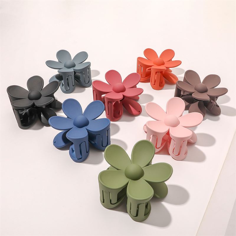 Eight SOHO Bloom Hair Claws, including the SOHO Bloom Hair Claw - Pink, are arranged in a semi-circle on a white surface. Each SOHO hair clip features a unique flower shape and comes in black, dark blue, blue, green, pink, brown, red, or orange.