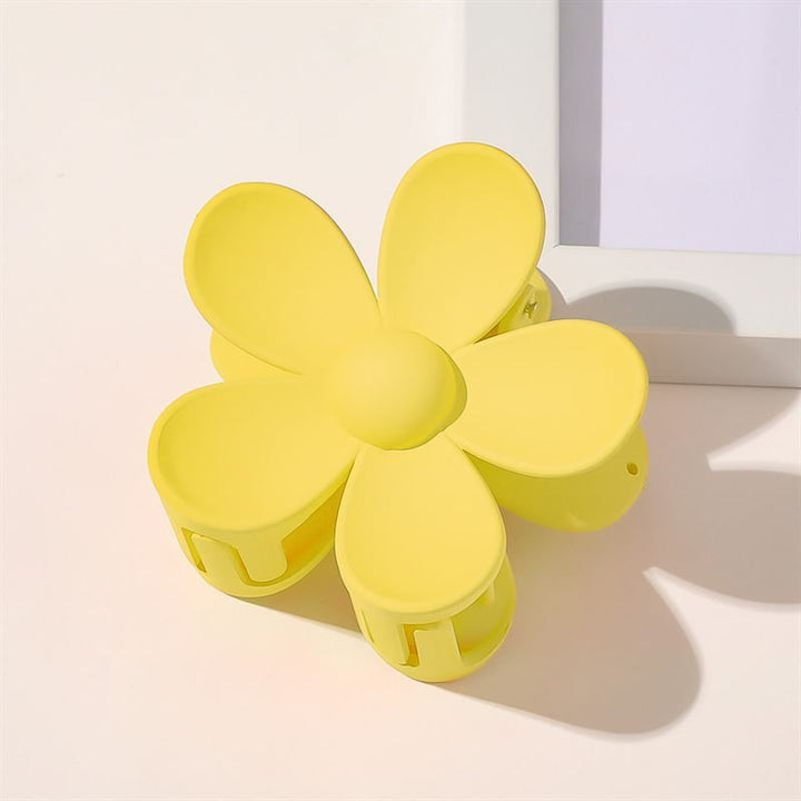 Die SOHO Bloom Hair Claw – Yellow, ein blumenförmiges Haaraccessoire von SOHO, liegt auf einer hellen Fläche in der Nähe der Ecke eines weißen Bilderrahmens.