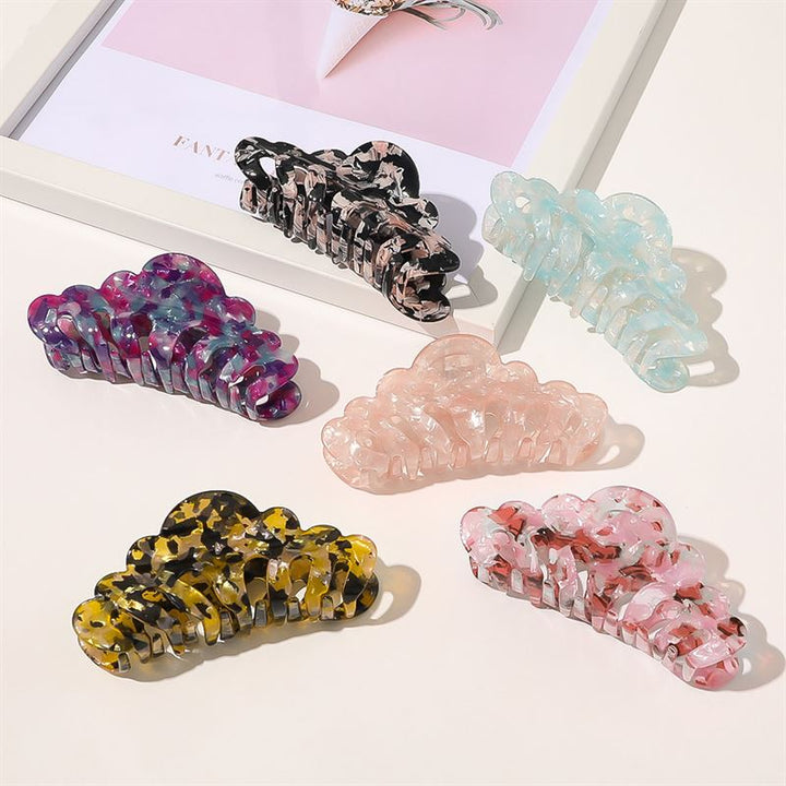 Five vibrant SOHO Callie Hair Claws in blush, purple, blue, pink, yellow, and black are arranged on a white surface. In the background, a framed photo highlights another large SOHO hair claw from the collection.