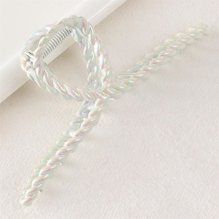 Die SOHO Caprina Hair Claw – White von SOHO verfügt über eine schillernde, glasierte Chromoberfläche und ein Design mit gedrehten Seilschleifen, die auf einer hellen, strukturierten Oberfläche ruhen.