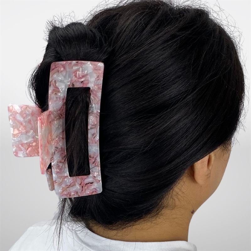 Eine Person mit dunklem Haar in einer tiefen Flechtfrisur, die mit der SOHO Clara Hair Claw – Light Coral befestigt ist. Die XL-Haarspange hält dickes Haar sicher fest. Sie trägt ein weißes Hemd und wird von hinten vor einem einfarbigen Hintergrund gezeigt.