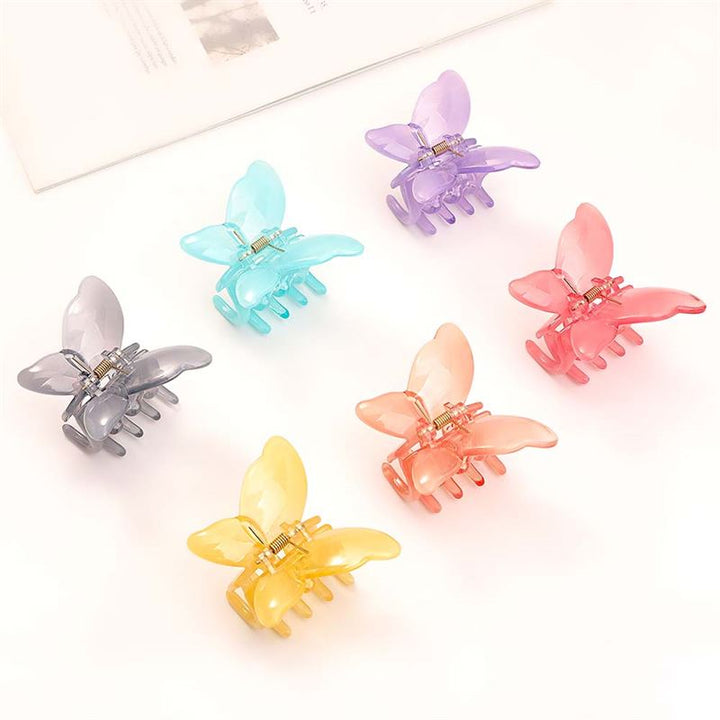 Die SOHO Ella Hair Claw – Arctic von SOHO besteht aus sechs pastellfarbenen, schmetterlingsförmigen Acetat-Clips – lila, blau, rosa, grau, orange und gelb –, die in einem Kreis auf einer weißen Fläche neben einem weißen Buch für niedliche Frisuren angeordnet sind.