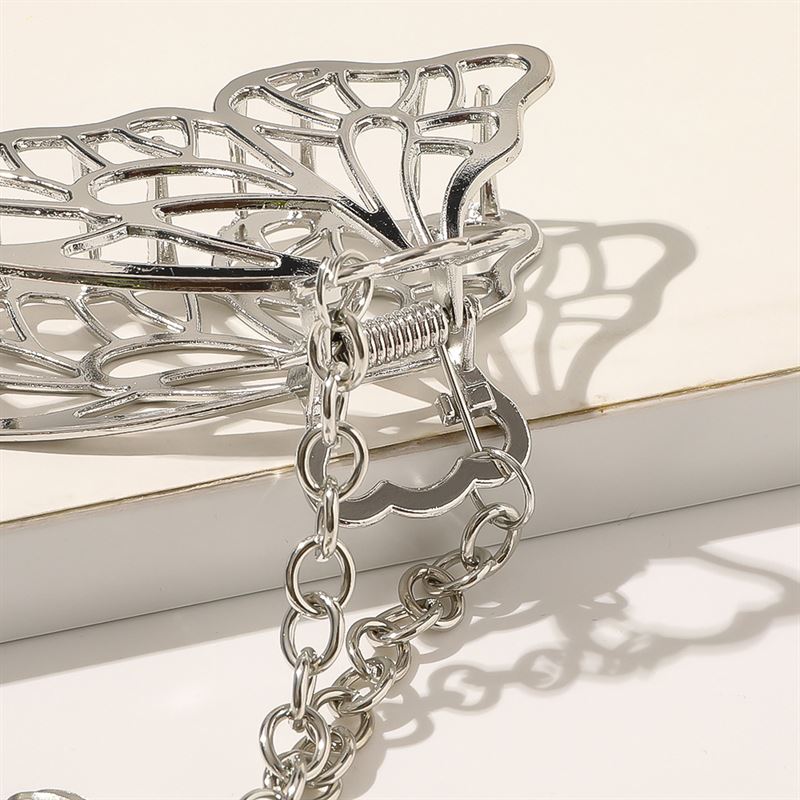 A close-up of the SOHO Emy Metal Hair Claw - Butterfly by SOHO, a shiny silver butterfly-shaped hair claw with an attached chain, resting on an off-white surface with soft shadows in the background.