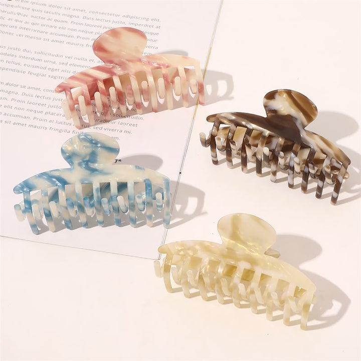 Die SOHO Gaja Hair Claw in Beige Marble sowie Accessoires in Rosa, Braun und Blau sind kunstvoll auf einer ebenen Fläche neben einem aufgeschlagenen Magazin arrangiert, während das Sonnenlicht sanfte Schatten auf diese elegante SOHO-Kollektion wirft.