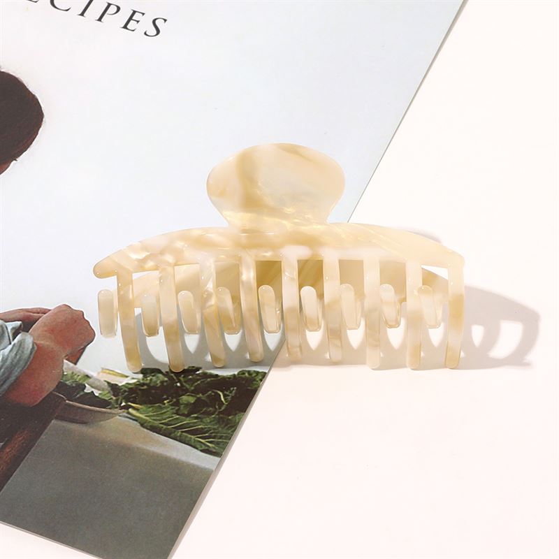 Die SOHO Gaja Hair Claw in Beige Marble liegt auf einem Magazin, ihr elegantes Marmordesign wird durch weiche Schatten hervorgehoben. Das Foto auf dem Magazin zeigt Grünpflanzen und eine Person, die das schicke SOHO-Accessoire ergänzen.