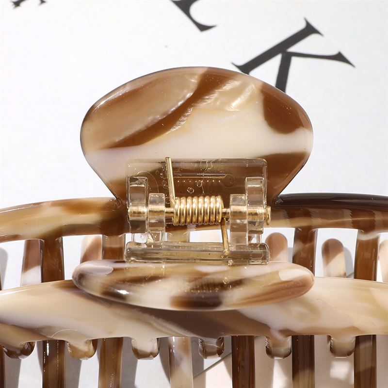 A close-up of the SOHO Gaja Hair Claw - Brown Marble by SOHO, highlighting its spring mechanism and curved prongs against a blurred background with partial black text.