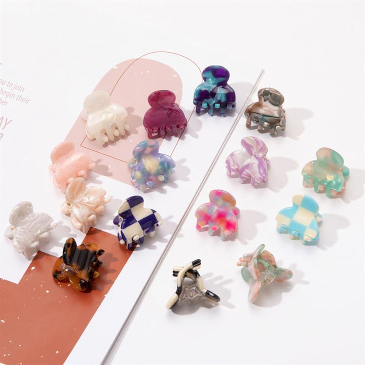 Die SOHO Hara Mini Hair Claw in Cloud Pink von SOHO wird zusammen mit anderen bunten Mini-Haarklammern auf einer weißen Fläche und einem Notizbuch präsentiert, wodurch ihre einzigartigen Muster und Formen besonders gut zur Geltung kommen.