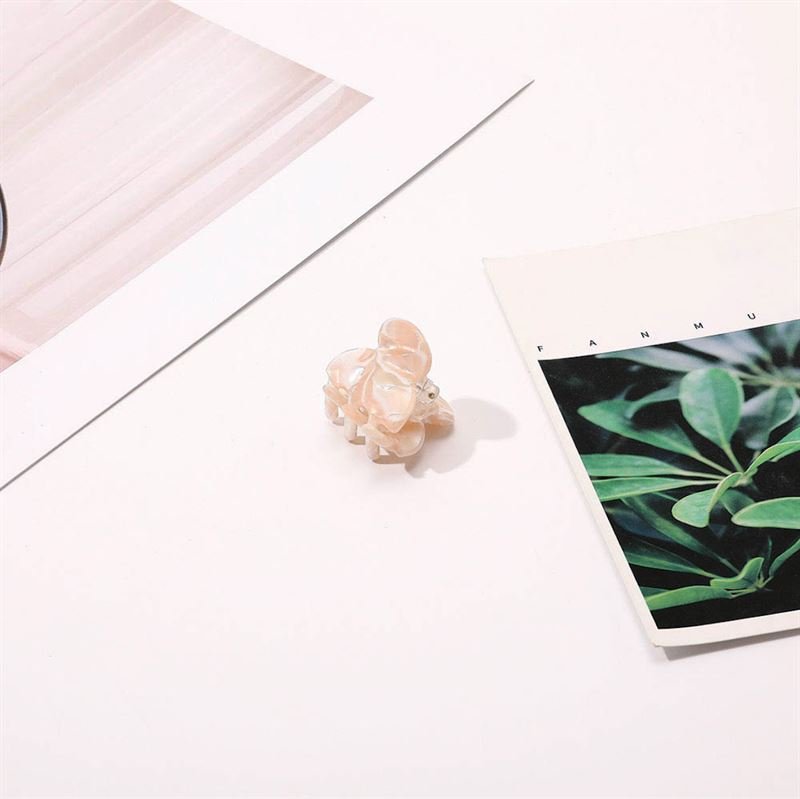 Eine kleine, hellrosa Elefantenfigur sitzt auf einer weißen Fläche neben der SOHO Hara Mini Hair Claw – Cloud Pink von SOHO, zwischen zwei Papieren – eines mit einem Foto einer grünen Blattpflanze und das andere mit abstrakten hellrosa Linien.