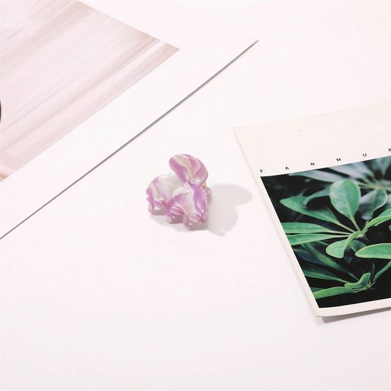 Die SOHO Hara Mini Hair Claw in Periwinkle – ein kleines, schmetterlingsförmiges Haaraccessoire mit marmorierten violetten und weißen Farbtönen – liegt auf einer weißen Fläche neben zwei Blättern Papier: eines mit grünen Blättern, das andere gestreift.