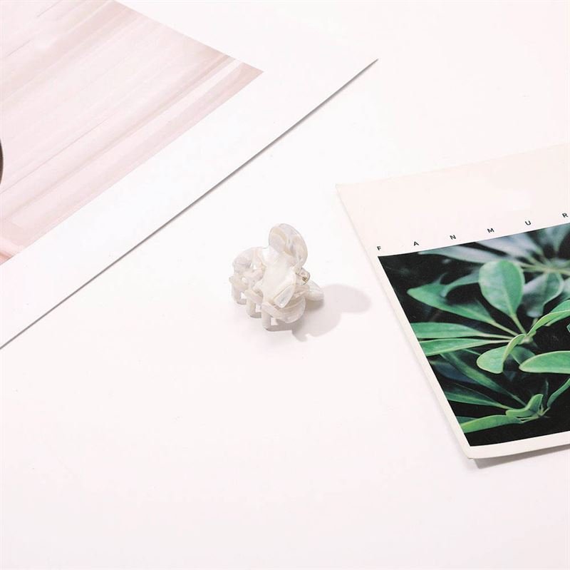 Die SOHO Hara Mini Hair Claw – Porcelain von SOHO, eine kleine weiße, durchscheinende Klammer, ruht auf einer weißen Oberfläche zwischen Pflanzen und abstrakten Papierakzenten und bietet einen sicheren Halt für das Haar und einen stilvollen Touch.