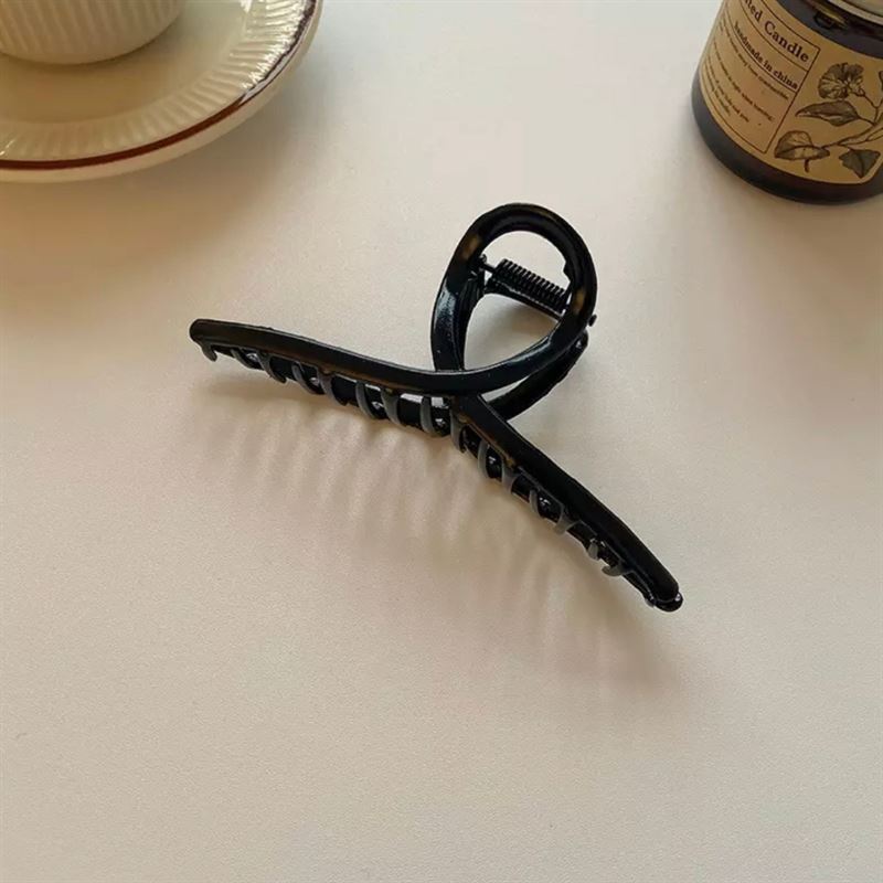 Die SOHO Iva Metal Hair Claw in Schwarz von SOHO wird auf einer weißen Fläche neben einer Teetasse, einer Untertasse und einem braunen Glaskerzenglas präsentiert – ideal, um unordentliche Dutts zu fixieren oder Ihrem Haar einen schicken Stil zu verleihen.