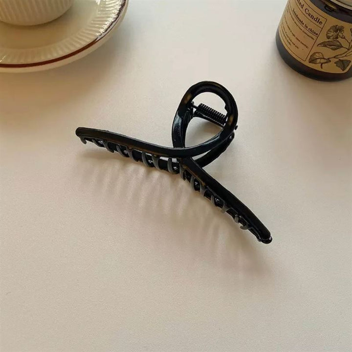 Die SOHO Iva Metal Hair Claw in Schwarz von SOHO wird auf einer weißen Fläche neben einer Teetasse, einer Untertasse und einem braunen Glaskerzenglas präsentiert – ideal, um unordentliche Dutts zu fixieren oder Ihrem Haar einen schicken Stil zu verleihen.