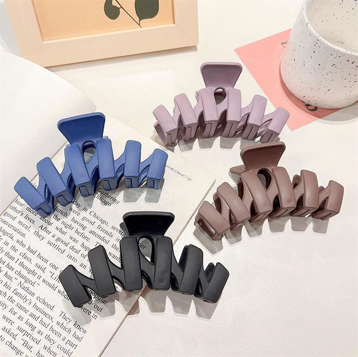 The SOHO Just Hair Claw in Black by SOHO, a stylish matte large hair clip, is displayed on an open book beside a white speckled mug and a peach frame with a plant illustration.