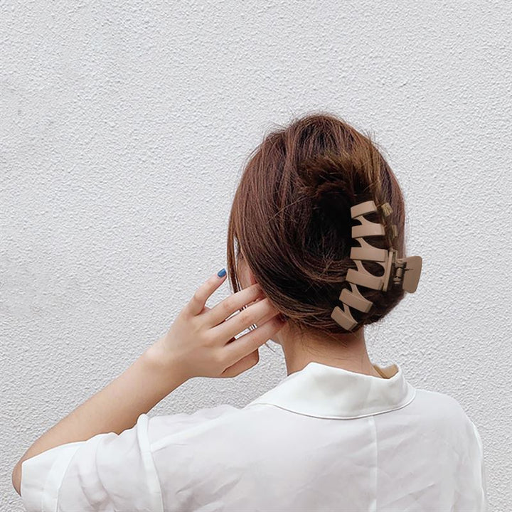 A person with medium brown hair in a bun, secured with the SOHO Just Hair Claw - Black by SOHO, stands facing a white textured wall, wearing a white collared shirt and touching their hair with their left hand.