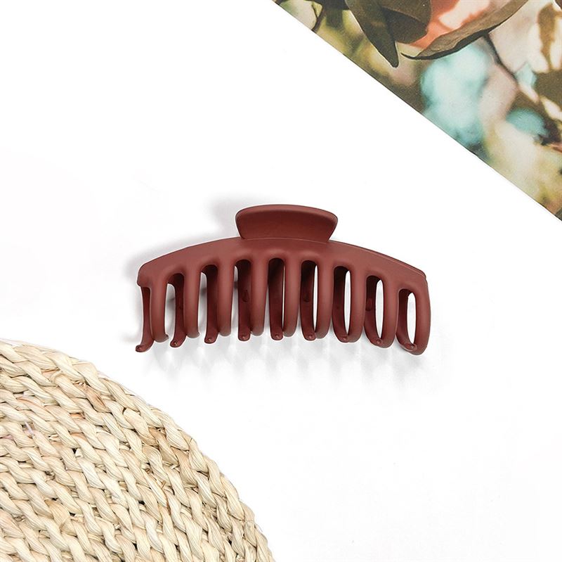 The SOHO Kana Hair Claw in Bark Brown, a matte wide-tooth clip for all hair types, sits on a white surface next to a woven mat and the corner of a colorful magazine.