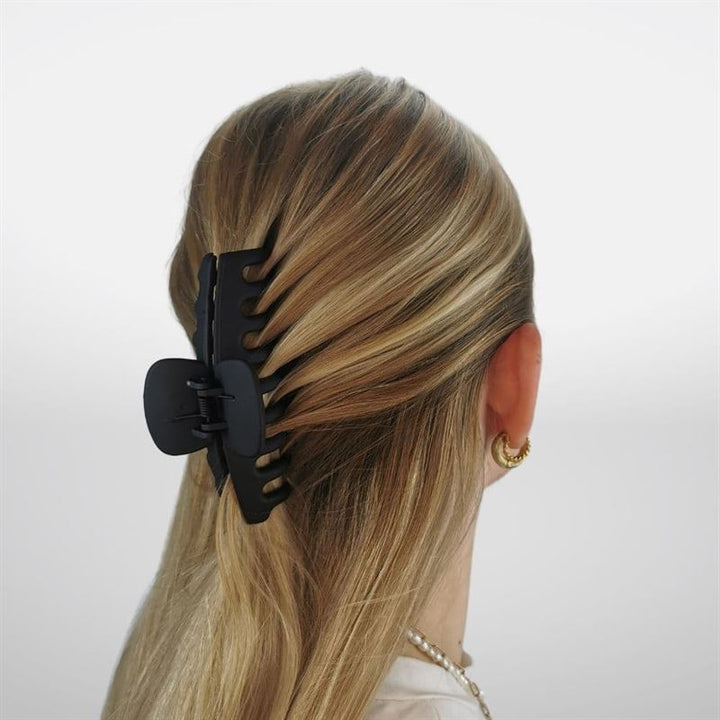 A woman with long, blonde hair secures her hair back using the SOHO Kana Hair Claw - Black. Shown from behind, she wears gold hoop earrings and a white top against a light background. Ideal for all hair types. Brand: SOHO.