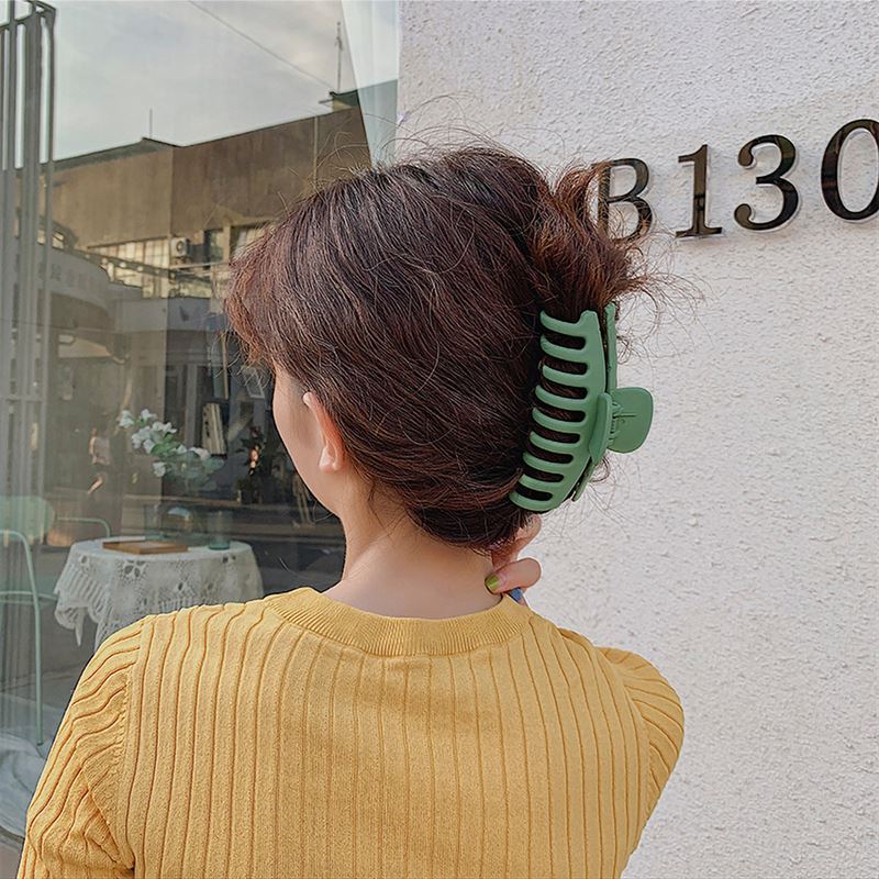 A person with wavy brown hair, in a mustard yellow top, stands near a white wall marked B130. Their hair is styled in a loose updo with the SOHO Kana Hair Claw - Peach Beige, known for its strong grip and suitability for all hair types.
