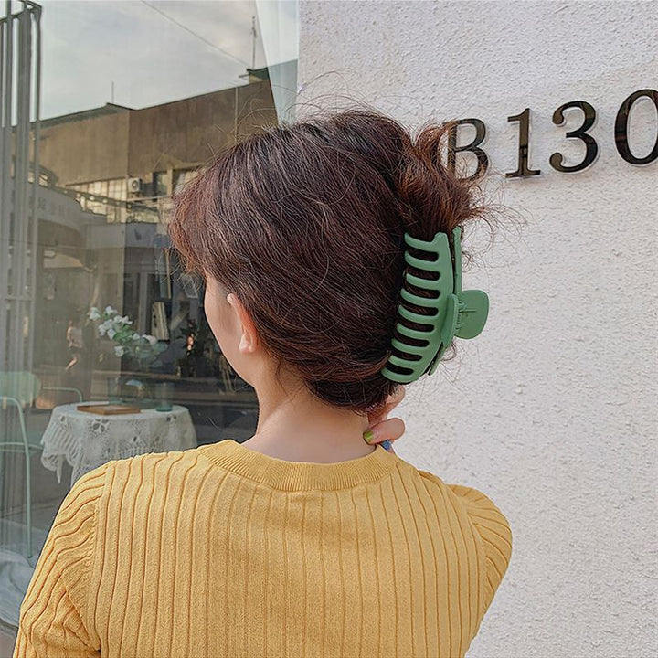 A person with wavy brown hair, in a mustard yellow top, stands near a white wall marked B130. Their hair is styled in a loose updo with the SOHO Kana Hair Claw - Peach Beige, known for its strong grip and suitability for all hair types.