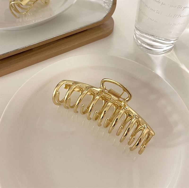 Die SOHO Kana Metal Hair Claw in Gold von SOHO, die für alle Haartypen geeignet ist, zeichnet sich durch einen starken Halt aus und befindet sich auf einer weißen Platte mit einem Spiegel und einem klaren Wasserglas im Hintergrund auf einer hellen Oberfläche.