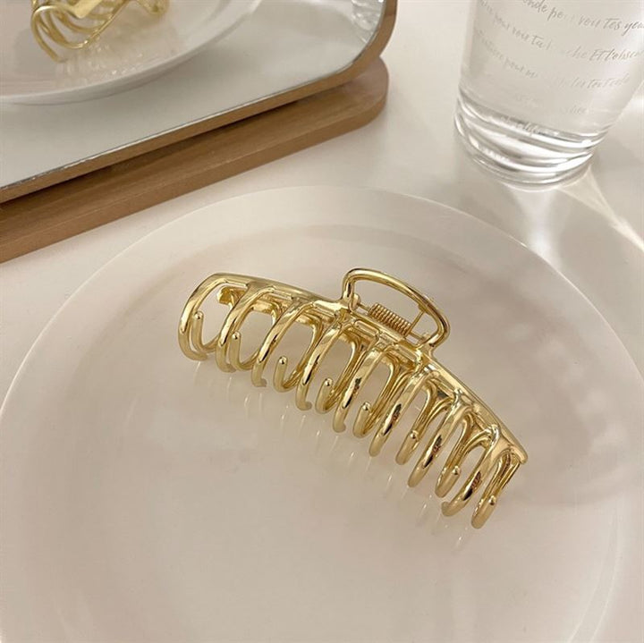 Die SOHO Kana Metal Hair Claw in Gold von SOHO, die für alle Haartypen geeignet ist, zeichnet sich durch einen starken Halt aus und befindet sich auf einer weißen Platte mit einem Spiegel und einem klaren Wasserglas im Hintergrund auf einer hellen Oberfläche.