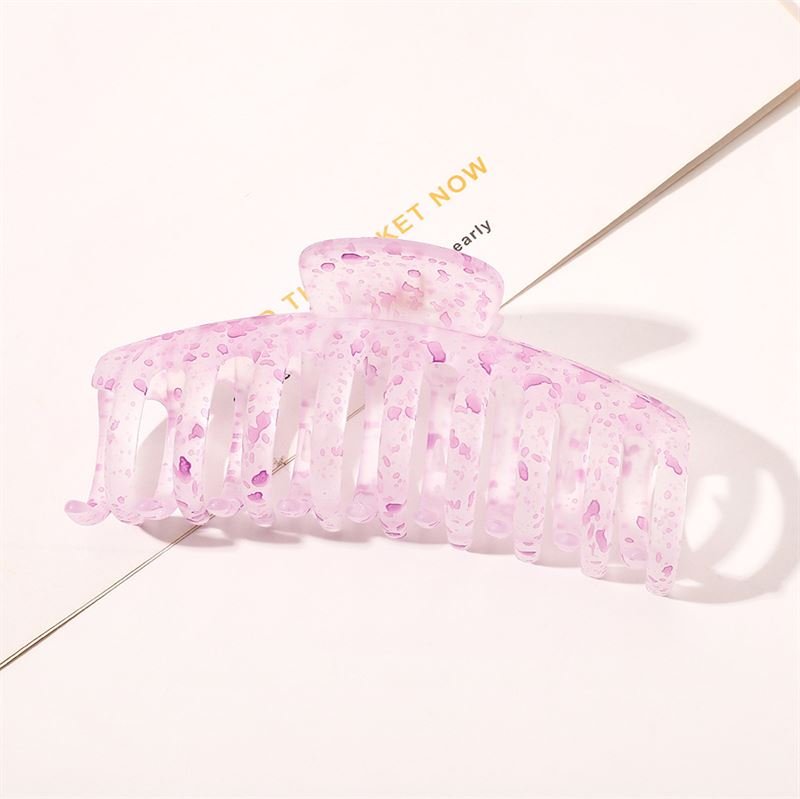 Die SOHO Kana Splash Hair Claw – Purple von SOHO hat ein durchscheinendes rosa Design mit violetten Sprenkeln und wird auf einer weißen Oberfläche mit darunterliegendem Text abgebildet. Diese Klammer bietet einen starken Halt und eignet sich für alle Haartypen.