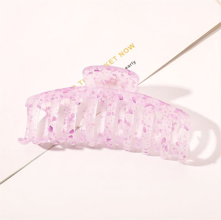 Die SOHO Kana Splash Hair Claw – Purple von SOHO hat ein durchscheinendes rosa Design mit violetten Sprenkeln und wird auf einer weißen Oberfläche mit darunterliegendem Text abgebildet. Diese Klammer bietet einen starken Halt und eignet sich für alle Haartypen.