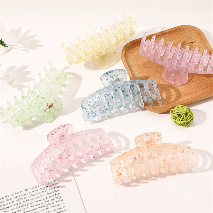 Die SOHO Kana Splash Hair Claw in Lila zeichnet sich durch ein pastellfarbenes Design und einen starken Halt für alle Haartypen aus. Präsentiert auf einer weißen Fläche mit einem Holztablett, einer grünen Dekokugel und einer aufgeschlagenen Zeitschrift in der Nähe.