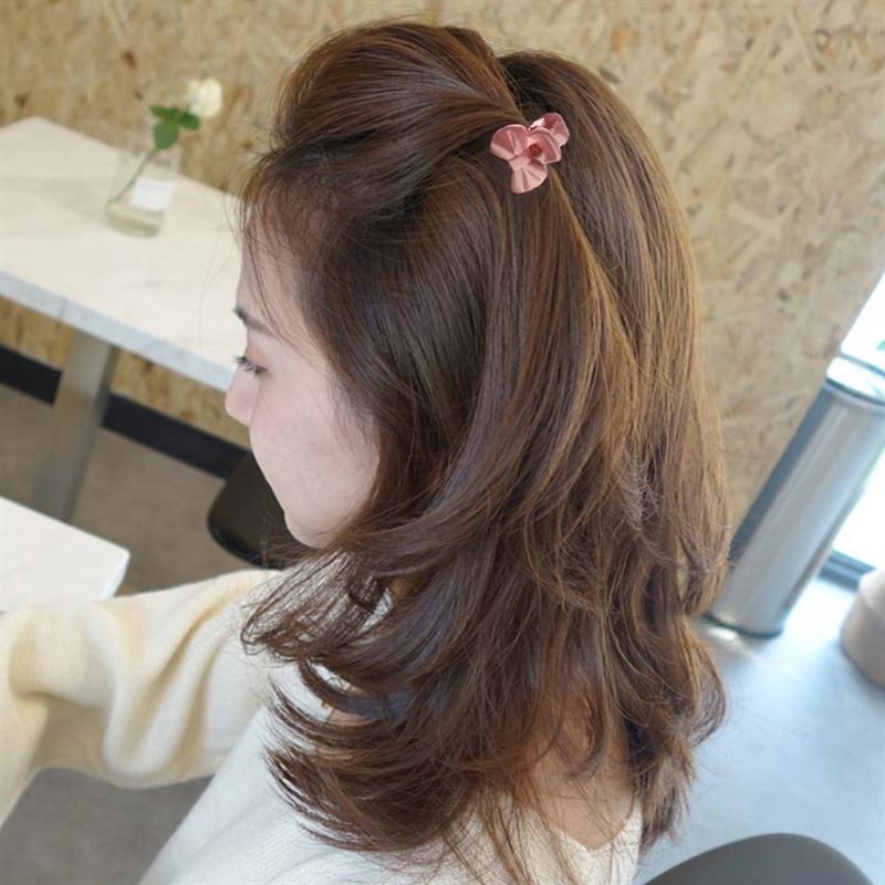 Eine Frau mit mittellangem braunem Haar sitzt an einem Tisch und trägt einen cremefarbenen Pullover. Ihr Haar ist mit SOHO Lala Mini Hair Claws – Flowers von SOHO gestylt. Im Hintergrund sind ein weißer Tisch, ein Stuhl und eine kleine Blumenvase zu sehen.