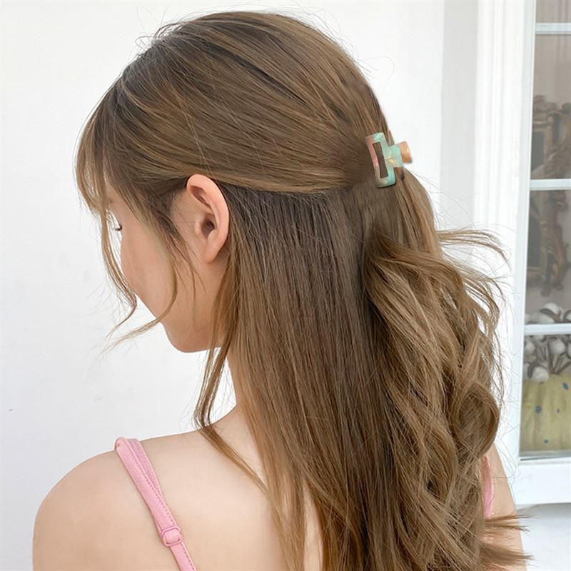 Eine Frau mit langen, welligen braunen Haaren trägt ein rosa Tanktop und befestigt ihr Haar mit der SOHO Leia Mini Hair Claw in Tortoise. Sie ist von hinten vor einem weißen Hintergrund zu sehen, wobei ein Teil eines Fensters sichtbar ist.