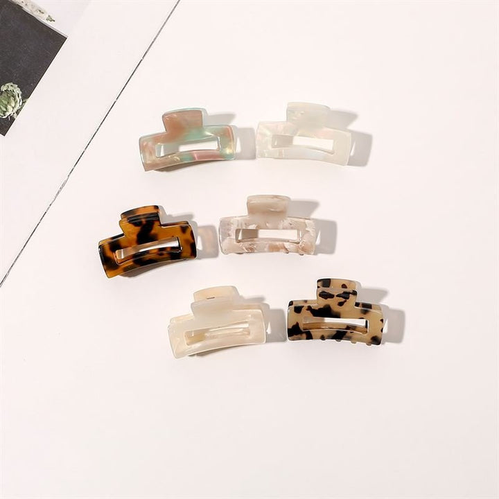 Sechs SOHO Leia Mini-Haarklammern in verschiedenen Farben und Mustern, darunter Schildpatt-, Creme- und Marmor-Designs, sind ordentlich auf einer hellen Oberfläche mit einer Zeitschrift im Blickfeld angeordnet – perfekt für stilvolle Frisuren.