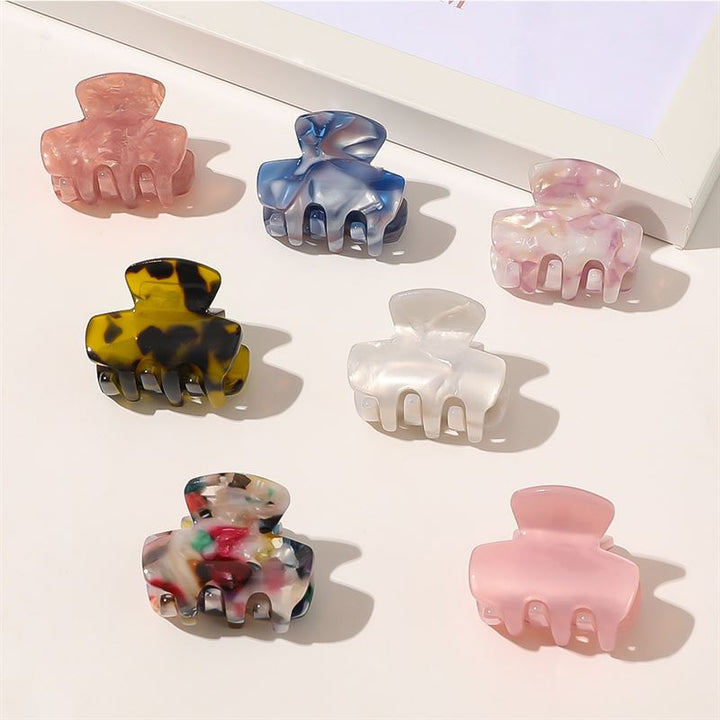 Sieben SOHO Loa Mini Hair Claws, darunter die Variante „White Marble”, sind ordentlich in Reihen auf einer hellen Fläche neben einem weißen Bilderrahmen angeordnet.