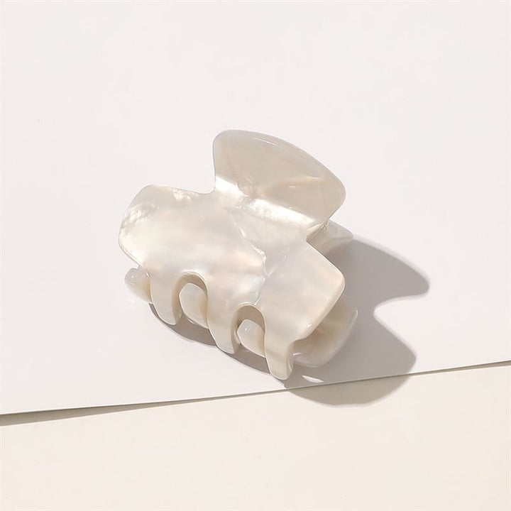 Die SOHO Loa Mini Hair Claw in White Marble verfügt über gebogene Zinken und einen perlmuttartigen Glanz und ruht auf einer weißen Oberfläche mit einem sanften Schatten.