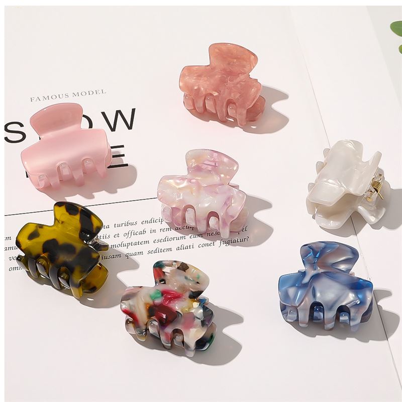 Sieben farbenfrohe SOHO-Haarklammern, darunter die SOHO Loa Mini Hair Claw in White Marble, sind kunstvoll in verschiedenen Formen und Mustern auf einer offenen Magazinseite unter heller, gleichmäßiger Beleuchtung angeordnet.