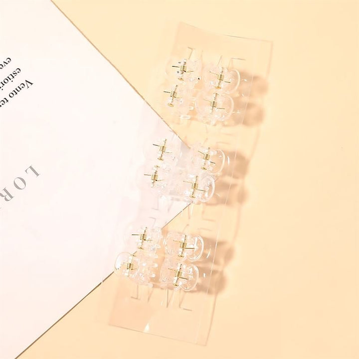 Zwei Paar klare Ohrringe mit goldenen Steckern sind auf einer pfirsichfarbenen Fläche neben einem weißen Blatt mit aufgedrucktem Text angeordnet, daneben SOHO Lucca Mini Hair Claws – Transparent von SOHO.