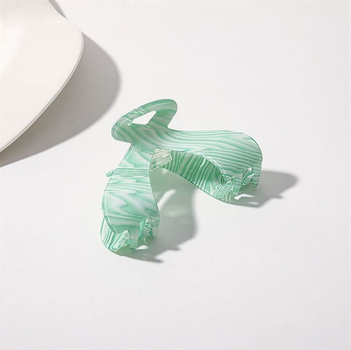 Die SOHO Malou Hair Claw – Green von SOHO liegt offen auf einer weißen Oberfläche, ihr marmoriertes grün-weißes Design wirft einen weichen Schatten. In der oberen linken Ecke erscheint ein weißes Objekt.