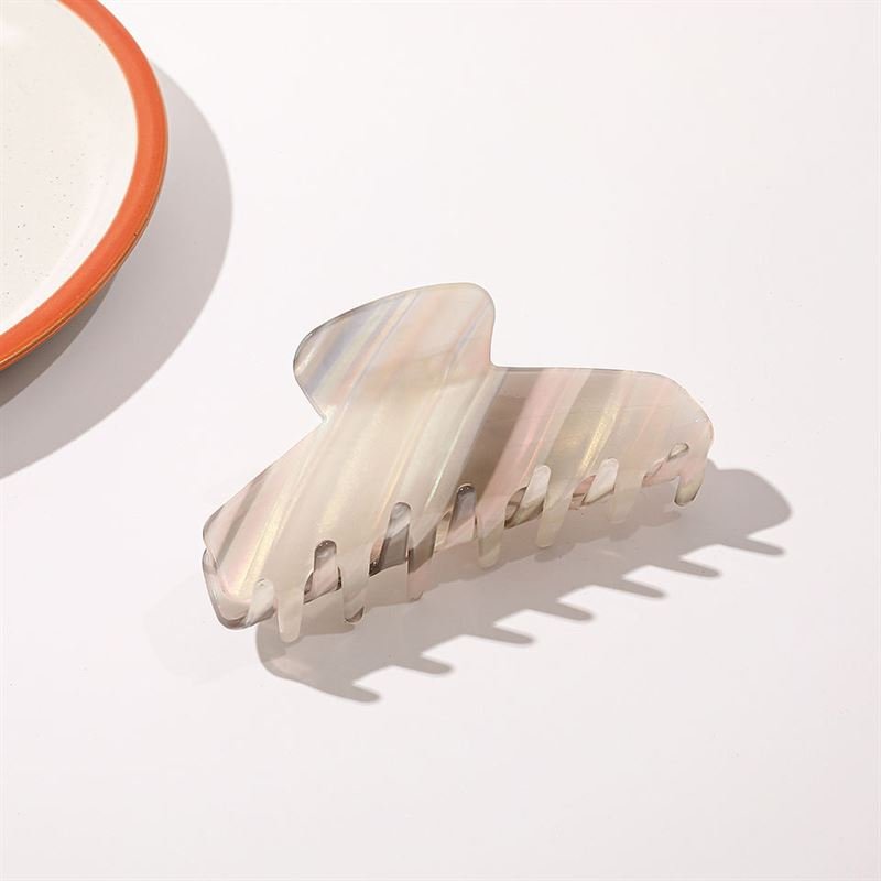 Die SOHO Mesa Hair Claw – Grey Rainbow von SOHO mit marmoriertem Perlglanzdesign liegt auf einer weißen Fläche nahe dem Rand eines weißen Tellers mit orangefarbenem Rand.