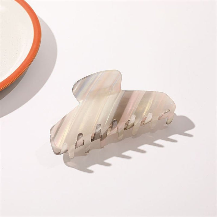 Die SOHO Mesa Hair Claw – Grey Rainbow von SOHO mit marmoriertem Perlglanzdesign liegt auf einer weißen Fläche nahe dem Rand eines weißen Tellers mit orangefarbenem Rand.