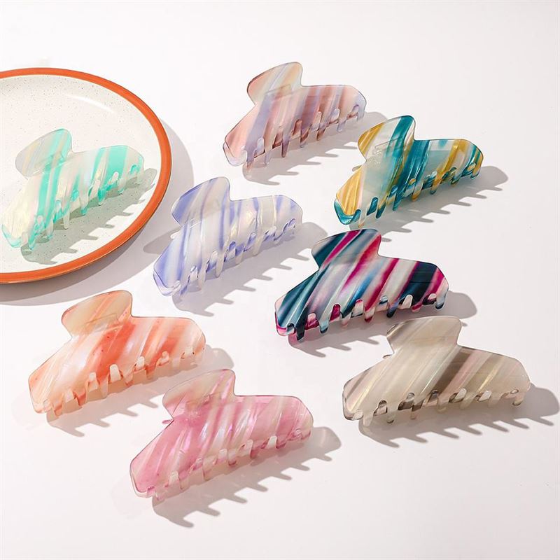 Sieben SOHO Mesa Hair Claw – Grey Rainbow Clips von SOHO werden auf einer weißen Fläche präsentiert. Ein kleiner runder Spiegel in der Ecke reflektiert zwei der Haarspangen, die jeweils mit einzigartigen pastellfarbenen oder leuchtenden grauen Regenbogenfarben verziert sind.