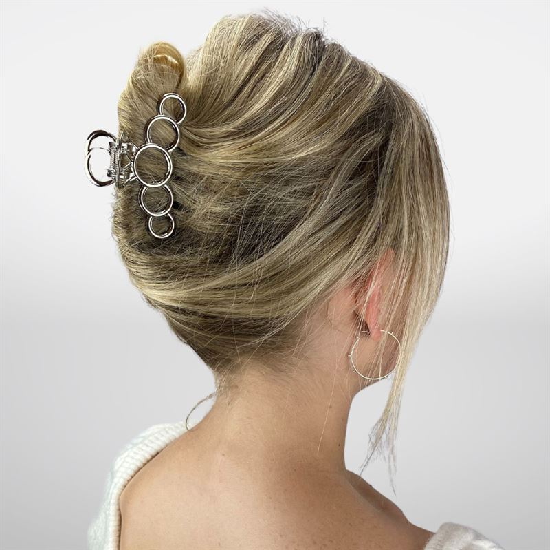 Eine Frau mit blondem Haar, das zu einem französischen Dutt gesteckt und mit der SOHO Metal Hair Claw in Silber fixiert ist – ideal für dickes Haar. Sie trägt einen cremefarbenen Pullover und silberne Creolen und ist von hinten vor einem weißen Hintergrund zu sehen.