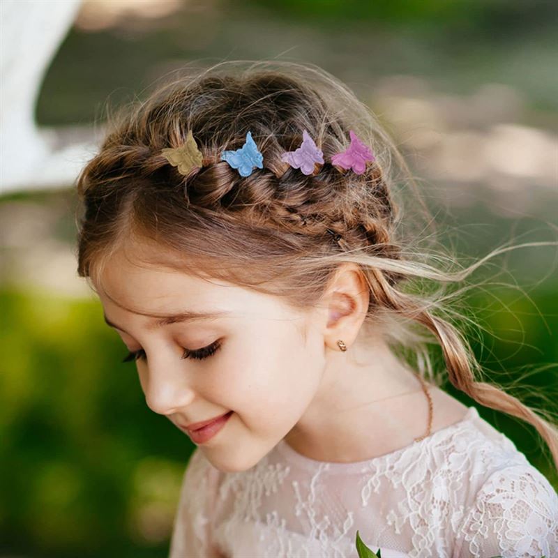 A young girl with braided hair adorned with SOHO Mini Butterfly Hair Claws (50 pcs, multicolour) smiles softly outdoors, wearing a light pink lace dress against a blurred green background.