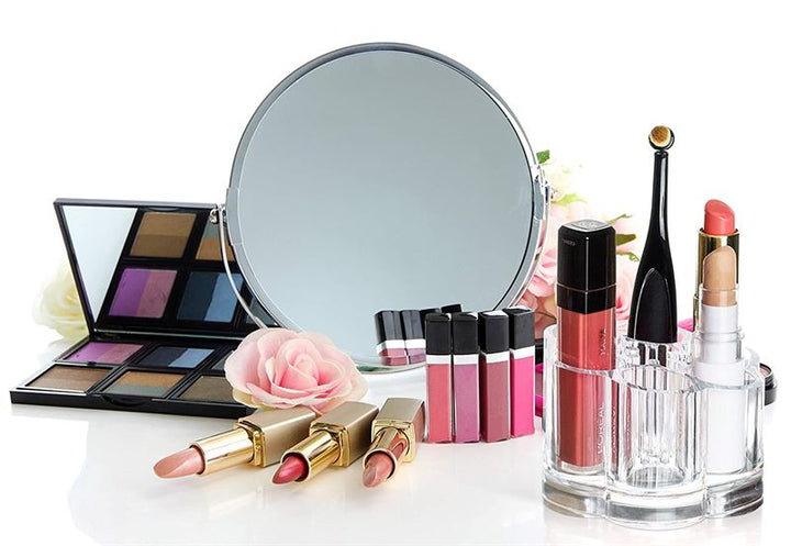 Der UNIQ Flower Makeup Acrylic Organiser – U301 von UNIQ präsentiert Lippenstifte, Lipgloss und Paletten zwischen rosa Kunstrosen und Pinseln vor einem runden Spiegel vor weißem Hintergrund.