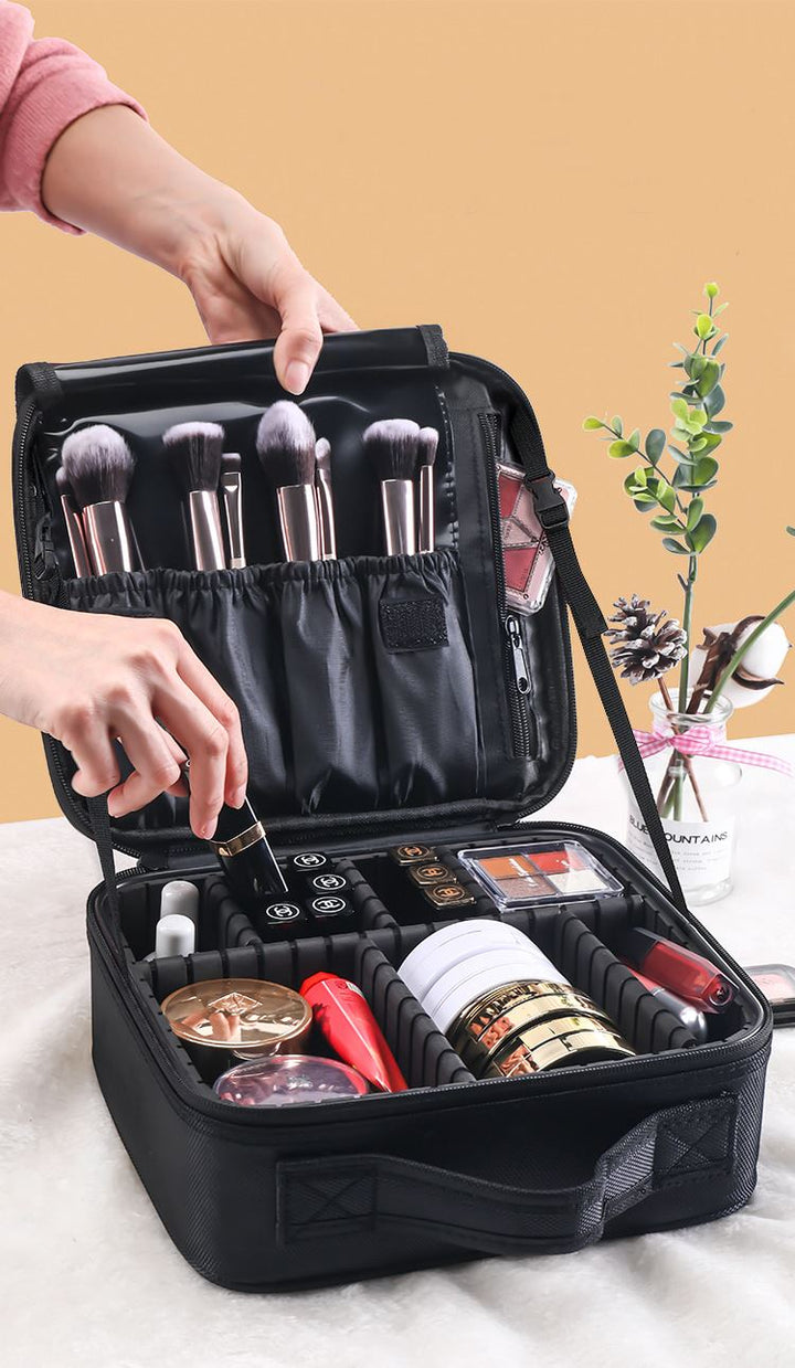 Jemand ordnet Make-up-Pinsel in der UNIQ Make-up-Reisetasche – einer verstellbaren schwarzen Kosmetiktasche von UNIQ –, die mit Kosmetika gefüllt ist, auf einem Tisch neben einer kleinen Topfpflanze.